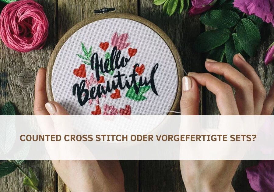Hand hält einen Stickrahmen mit gesticktem Schriftzug „Hello Beautiful“ und bunten Herzen auf weißem Stoff; umgeben von Garnrollen und Blumen; im Bild Text: „Counted Cross Stitch oder vorgefertigte Sets?