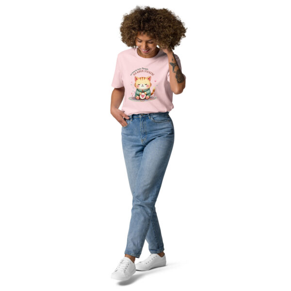 Frau mit lockigem Haar trägt ein Roséfarbenes T-Shirt mit Katzenmotiv und blauer Jeans. Auf dem T-Shirt ist eine lächelnde Katze mit einem Stickrahmen und Garn abgebildet. Darüber steht: ‚Ich brauche keine Therapie, ich kann sticken!‘ – Motiv von Vindalys. Die Frau blickt nach unten und lächelt, eine Hand in der Hosentasche, die andere am Haar.