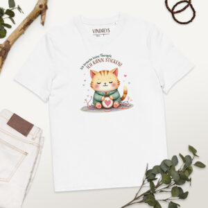 Weißes T-Shirt mit Katzenmotiv liegt flach auf einem hellen Untergrund. Das Motiv zeigt eine orangefarbene Katze mit einem Stickrahmen, darauf ein Herz. Um die Katze liegen bunte Fäden. Darüber steht der Text: ‚Ich brauche keine Therapie, ich kann sticken!‘ – Design von Vindalys. Daneben liegen eine helle Hose, grüne Blätter, ein Holzstück und ein Armband.